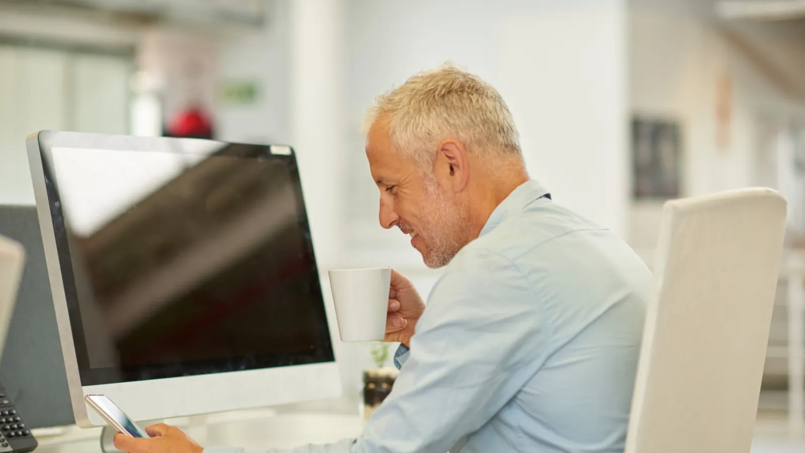 A man in an office sitting at a desk looking at his phone and drinking coffee