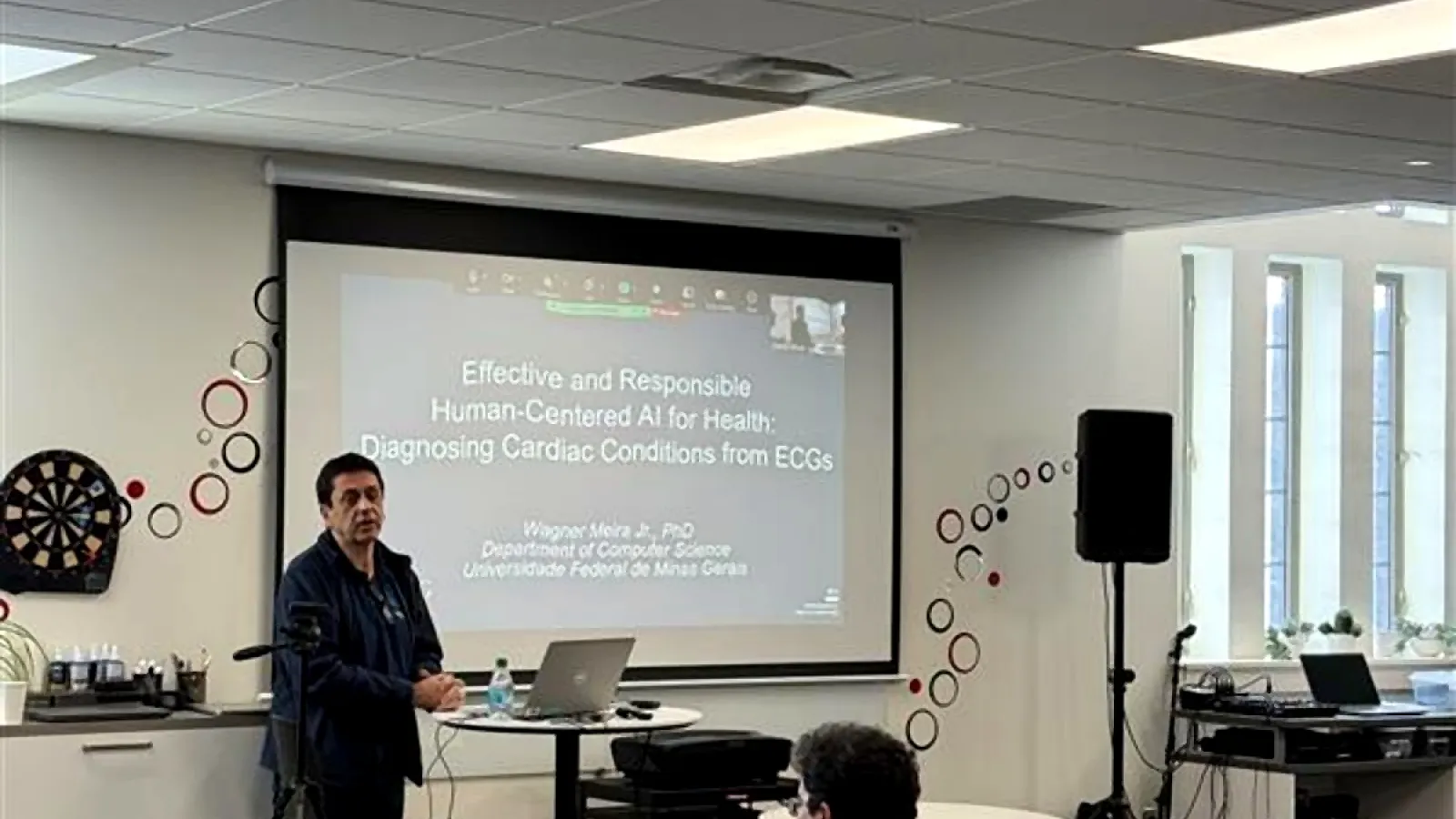 Wagner Meira Jr. presenting to an audience in Pomerene Hall with a projected slide on human-centered AI for health behind him.